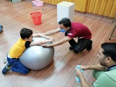 A young man rests his hands against a large vinyl stability ball, which a child is kneeling and hugging