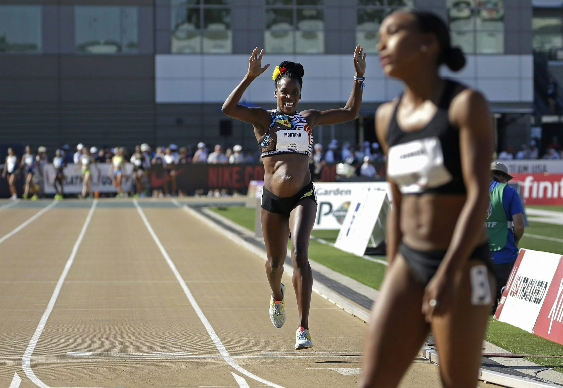 A pregnant Black woman in a track uniform smiles while raising her arms as she crosses a track finish line