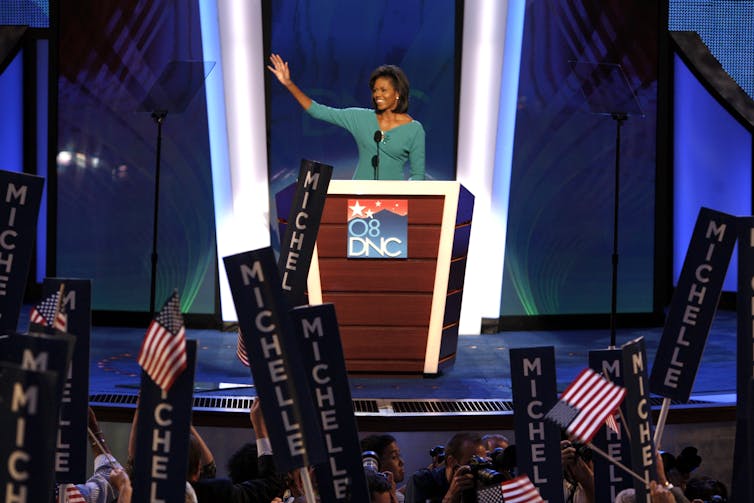A Black middle aged woman waves to the audience and wears a teal dress stands a podium that says '2008 DNC.' People in the audience hold posters that say 'Michelle.'