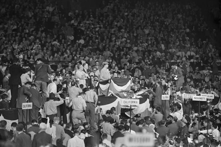 A black and white photo shows a woman standing at a podium, surrounded by people in a busy crowd.