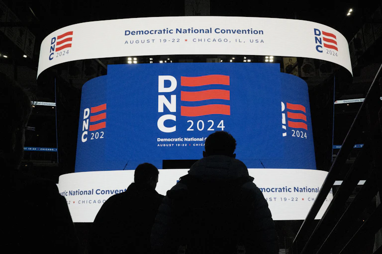 A large screen says DNC in white with red lines and there is a banner above it that says Democratic National Convention, August 19-22, Chicago, IL, USA