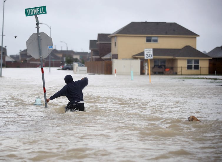 A person holds onto a spot sign post as he tries to wade through a waist-deep water in a flooded neighborhood of homes and pavement.