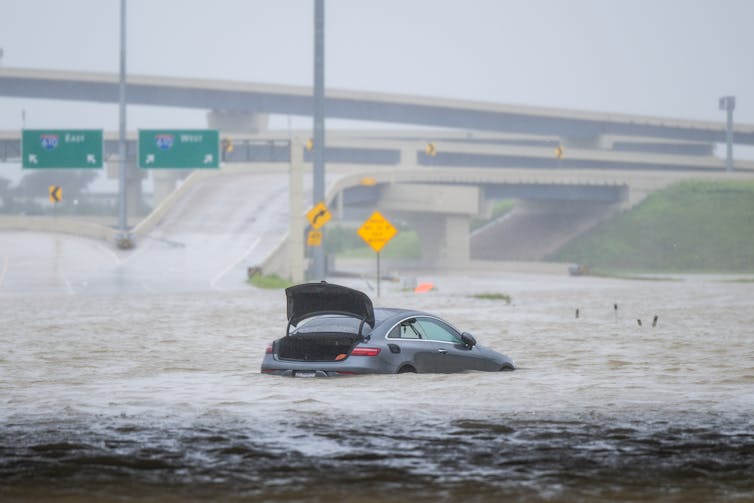 A car with is trunk lifted is stranded in floodwater up to its window with a flooded highway overpass in the background. Water flows along the pavement like rivers.