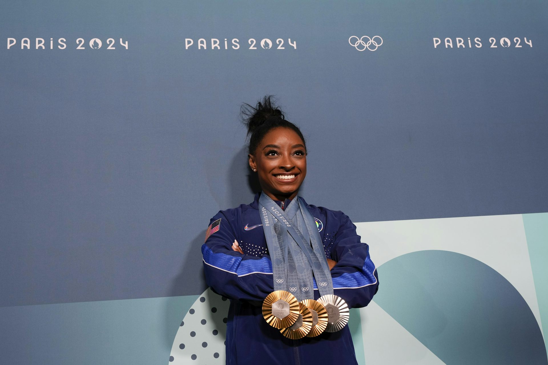 A female athlete wearing a track suit and several Olympic medals in front of a wall with 'Paris 2024' and the Olympic rings on it