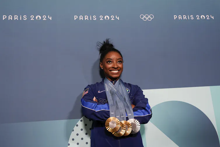 A female athlete wearing a track suit and several Olympic medals in front of a wall with 'Paris 2024' and the Olympic rings on it