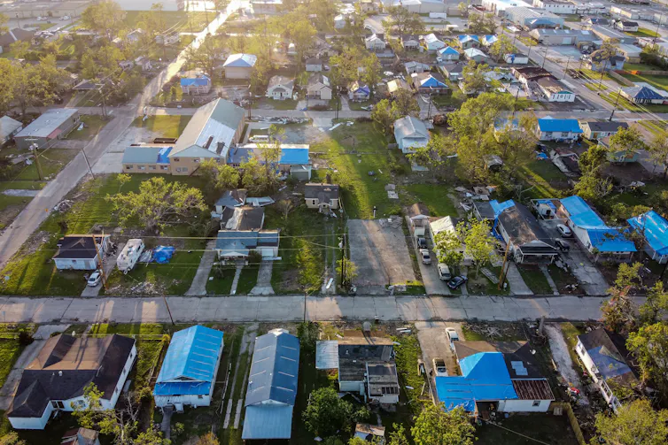 An aerial view of a neighborhood with most of the homes' roofs either missing or covered with a blue tarp.