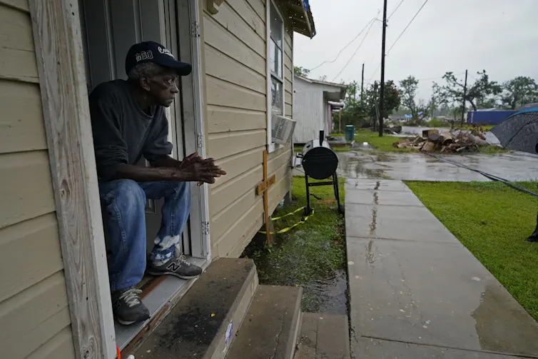 A man sits in the door of a house with a worried look on his face. Rain from Hurricane Delta is already falling.