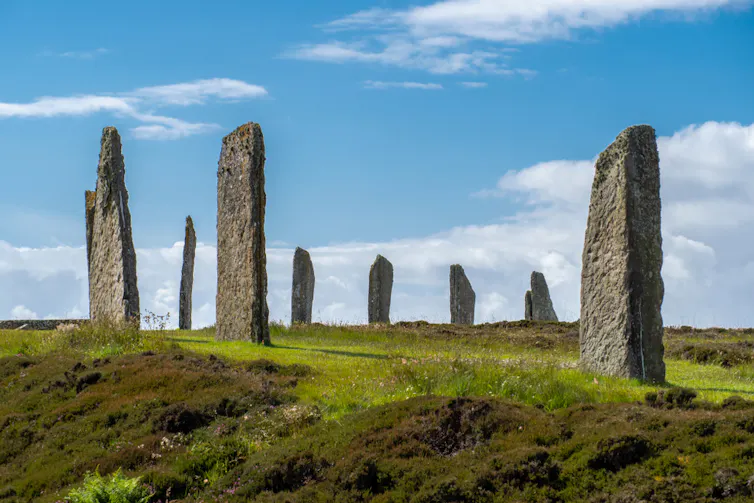Um círculo de pedras irregulares eretas no topo de uma colina sob um céu azul.