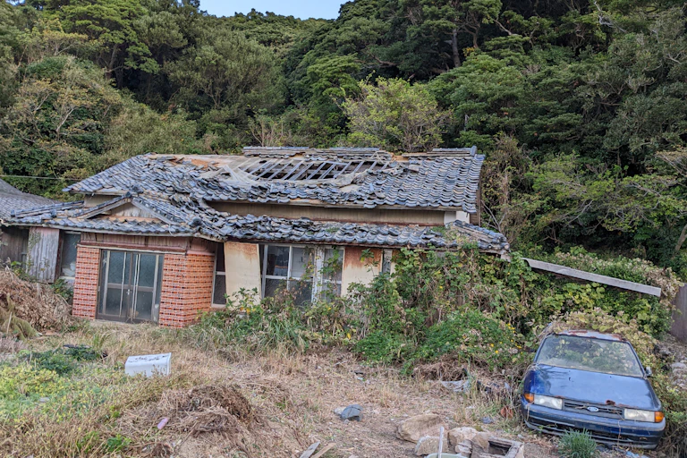 An abandoned and overgrown house on Naru Island with a blue car on the right.