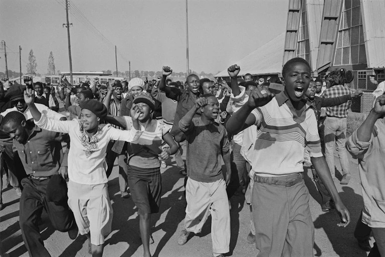 A black and white photo shows a large group of young protesters running along a road, fists and arms raised.