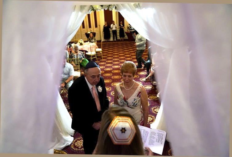 An older man in a skullcap and suit stands next to an older woman with short hair and a white dress in front of an officiant.