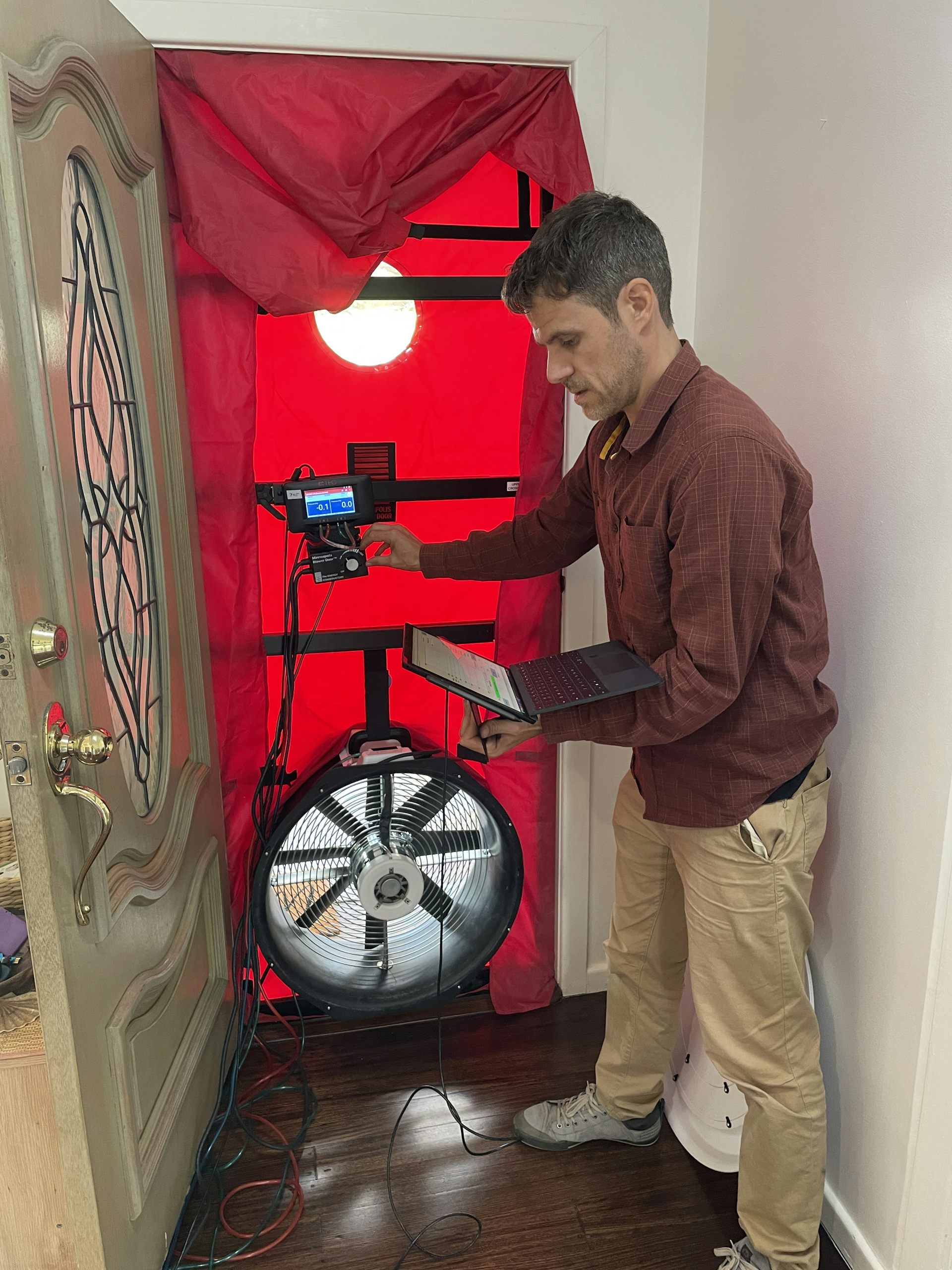 A person standing next to a blower door unit that is mounted in the front door of a house.