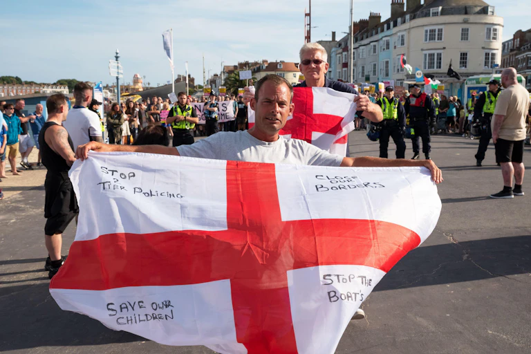 Two men wave English flags in front of a crowd. Writing on the flags reads 'stop two tier policing', 'close our borders', 'save our children' and 'stop the boats'