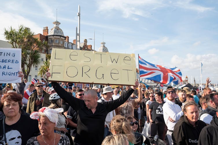 A man at a protest holding up a sign reading 'lest we forget'.
