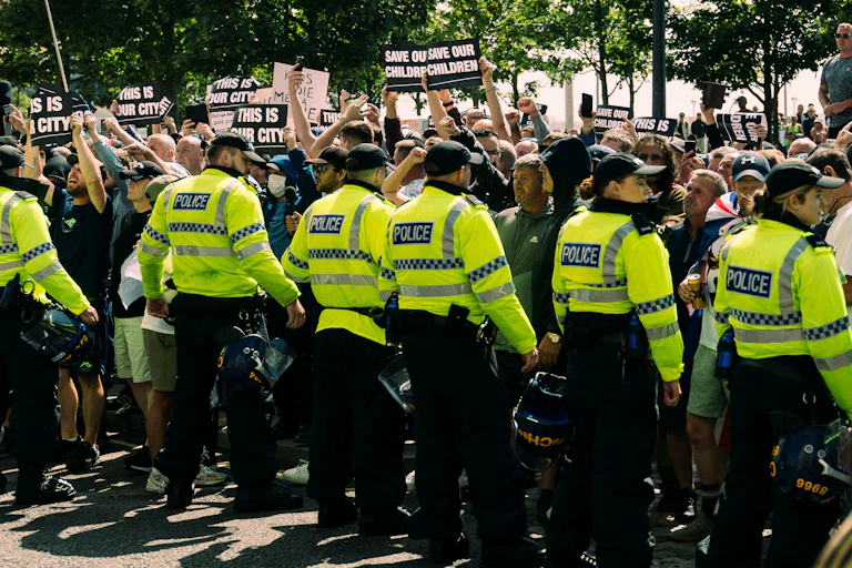 A line of police in hi-vis jackets facing a large crowd of mostly white men, holding signs that read 'save our children'.
