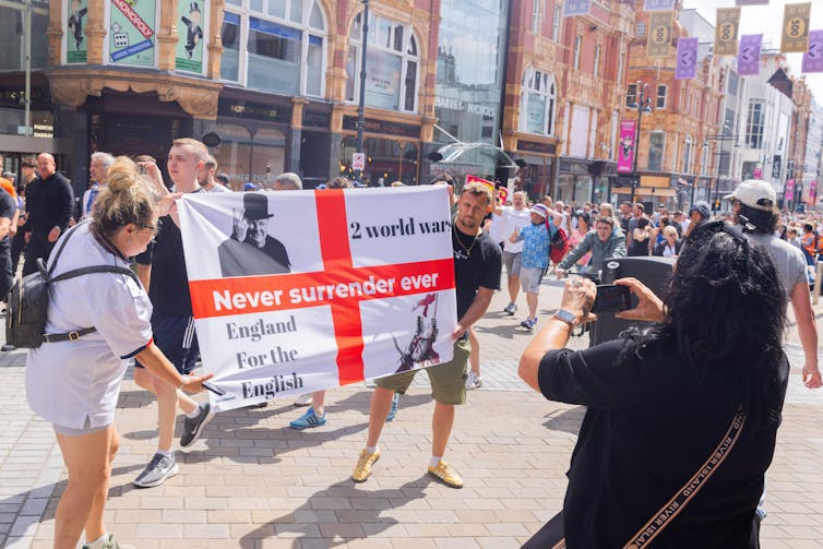 A man and a woman hold up an English flag with the words 'two world wars' and 'England for the English' while someone takes a photo.
