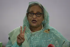 A woman wearing glasses in a pale green dress and head scarf flashes the victory symbol.