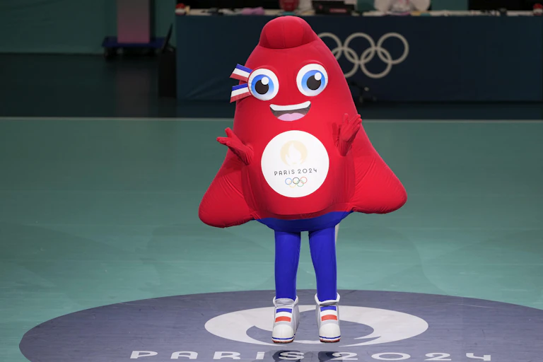 A red plush mascot with hands up in front of Olympic rings on an arena wall in the background.