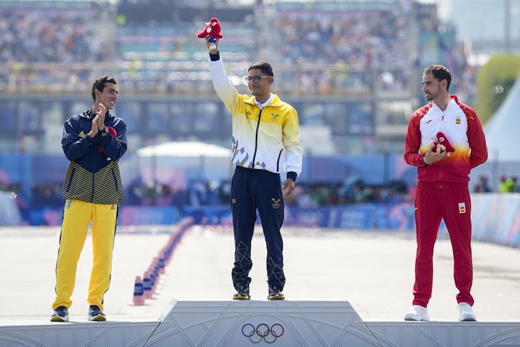 Athletes seen on a podium, the one in the centre lifting a red mascot.