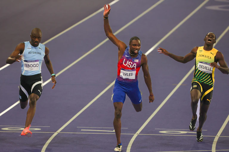 Three sprint runners on the track at the Paris Olympics, one with his hand in the air as he wins.