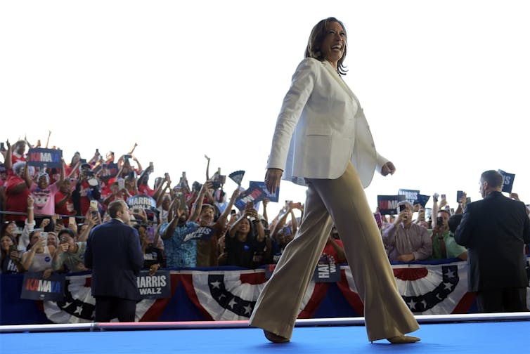 A dark-haired woman in a cream colour jacket and wide-legged beige pants smiles as she walks across a stage.