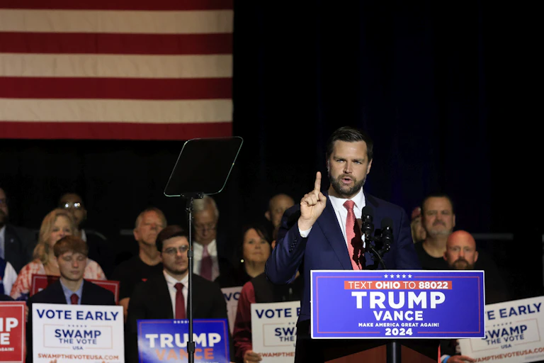 Young bearded man wearing blue suit jacket speaks at a lectern flanked by a crowd of people holding signs.