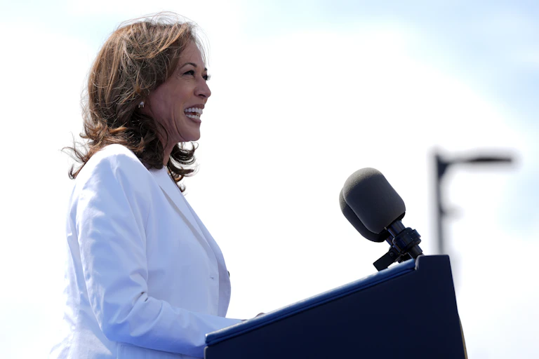 A dark-haired woman in a white jacket smiles as she stands behind a lectern.