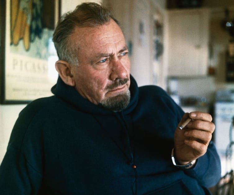 Older man with graying hair and a goatee poses while holding a cigarillo.