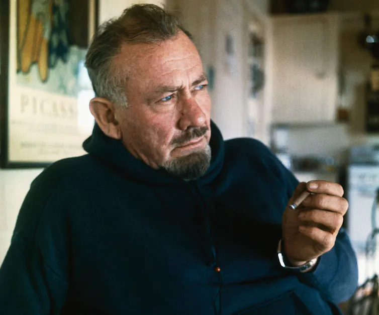 Older man with graying hair and a goatee poses while holding a cigarillo.
