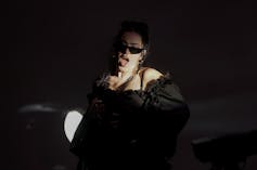 A woman on a darkly lit stage and wearing sunglasses sings into a microphone.