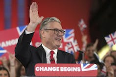A man with grey hair and glasses waves standing behind a lectern that has a Change Begins banner on it.