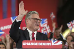 A man with grey hair and glasses waves standing behind a lectern that has a Change Begins banner on it.