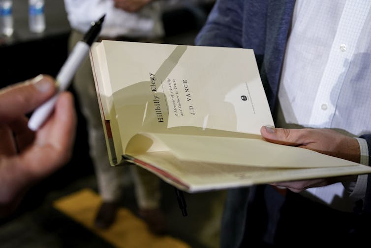 A man holds an open book as a hand presents a pen so the book can be signed.