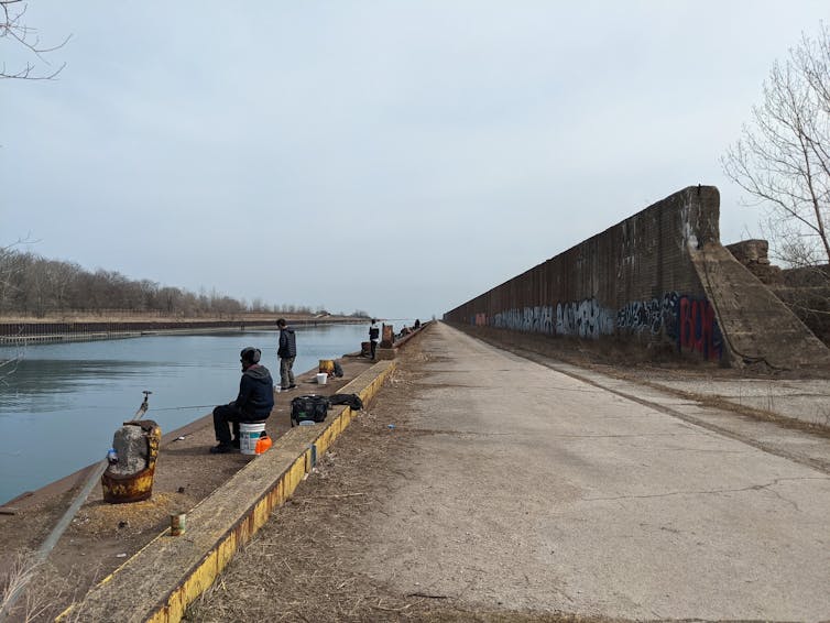 People sit on buckets with fishing poles on a chilly fall day. a large cement wall is behind them. The river is in front of them with Lake Michigan in the background.