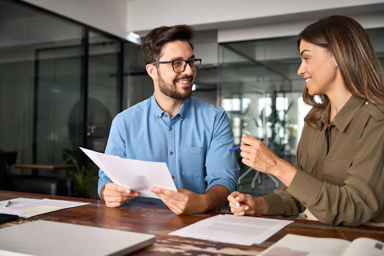 A man and a woman sit at a table in a workplace, having a discussion.