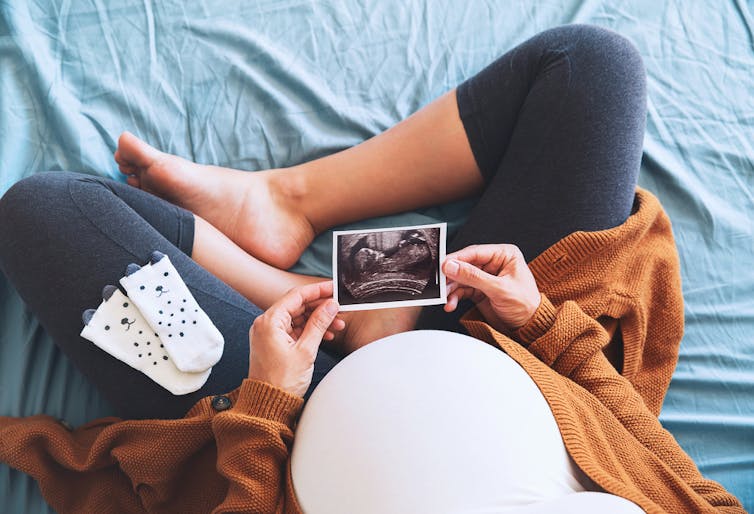 Pregnant woman sitting cross-legged on bed, seen from above, holding ultrasound image