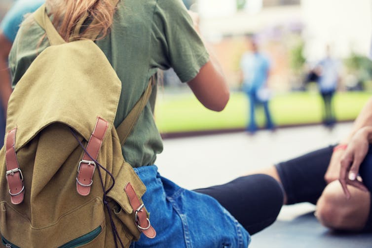 A young woman in a shirt and leggings, wearing a canvas backpack, sits on the ground.