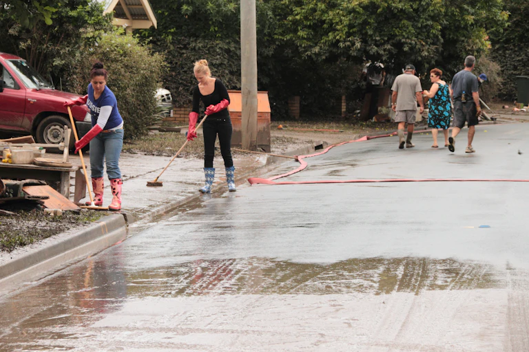Two women wearing rubber boots and gloves sweep water and mud from driveways in Fairfield, Brisbane after flooding. Three other people walk past.