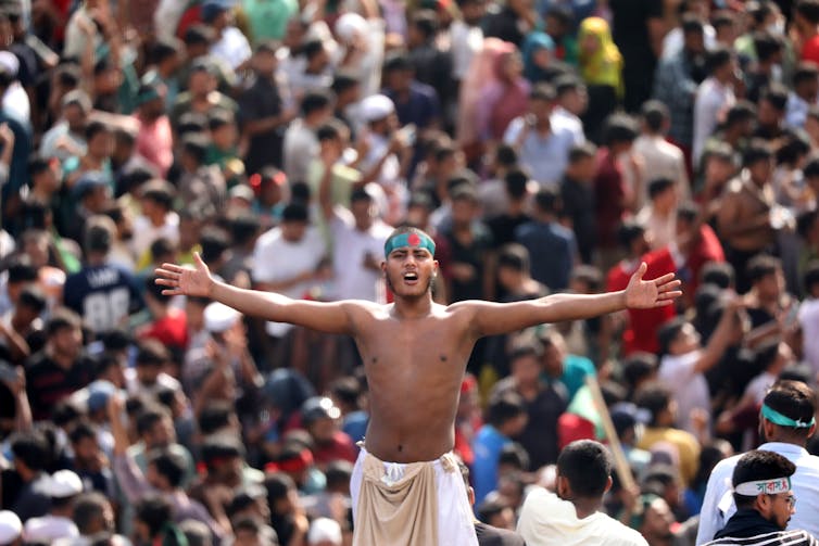 A Bangladeshi man standing with his arms outstretched above a crowd.