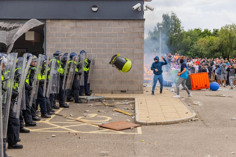 Riot police defending a burning building while a man throws a large object at them.
