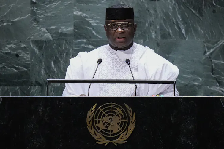 A middle-aged Black man in a white outfit, round glasses and a black kente hat speaks from behind a podium with the UN logo on it