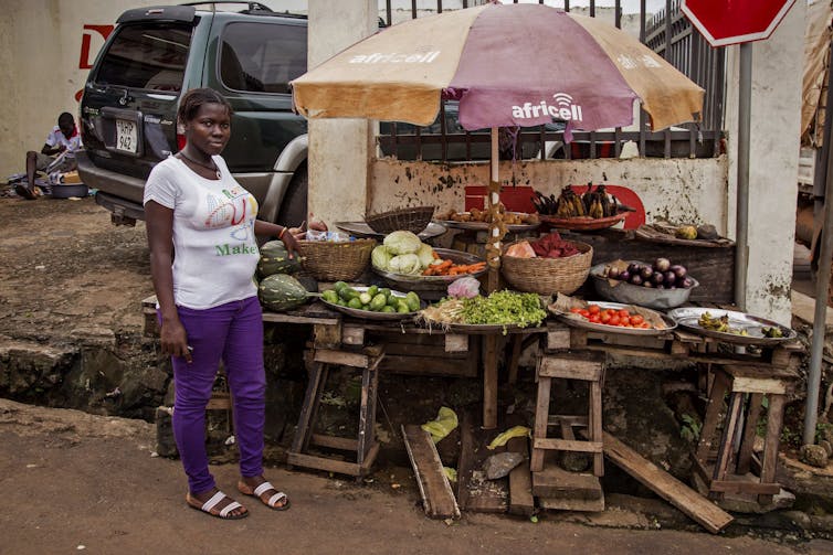 A Black woman in a white shirt and purple pants poses beside a display of fresh fruits and vegetables