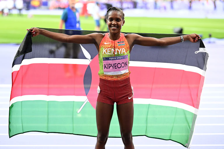 An African woman on a track in a stadium holds up a black, red and green flag behind her as she smiles for the camera.