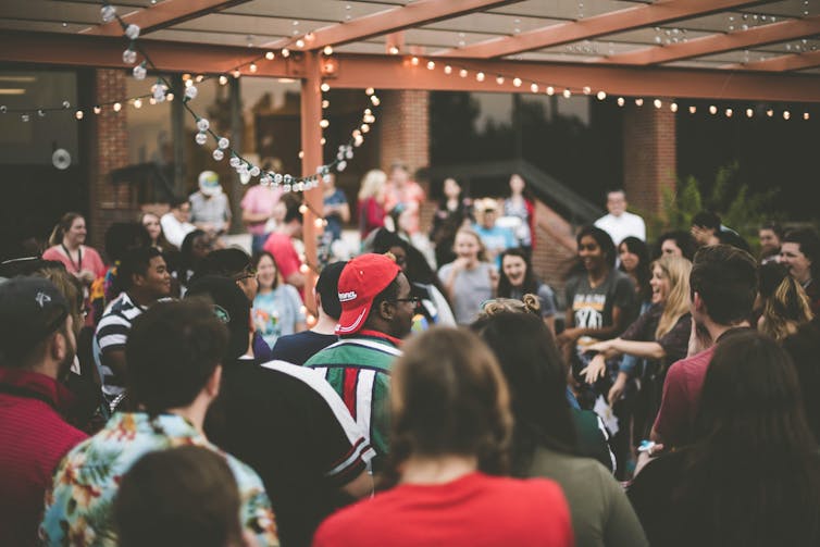 A group of young people stand outside in a crowd. Some are smiling and laughing. There are festoon lights above them.