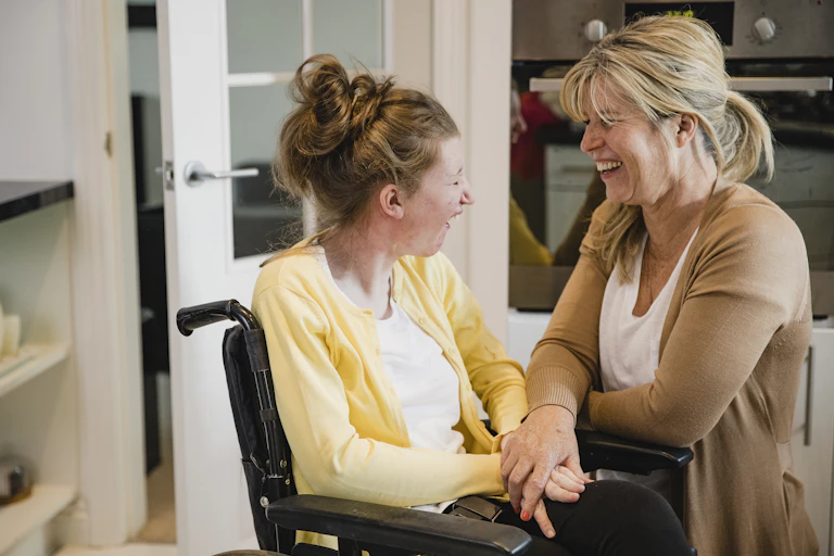 younger woman using wheelchair laughs with older woman in kitchen setting