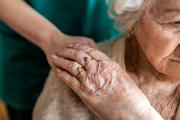 older woman is partially seen with someone hands on her shoulder in a reassuring way