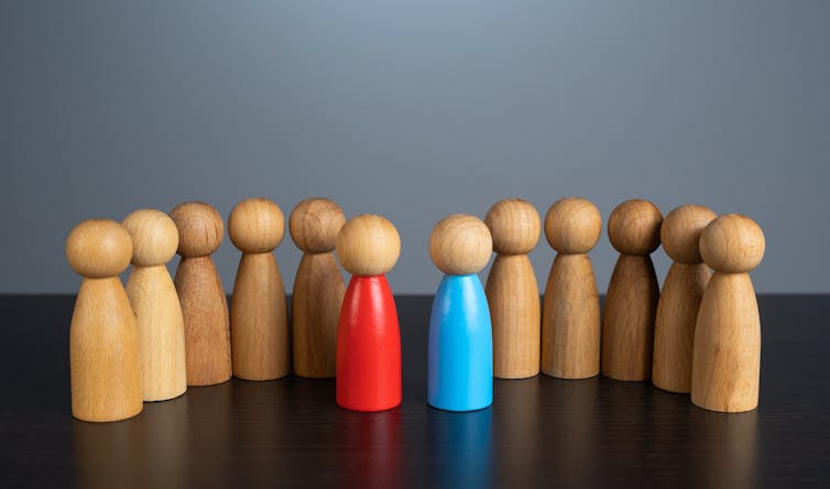 Two groups of small, simply wooden figures representing people, one group is led by a figure painted red, the other by a figure painted blue