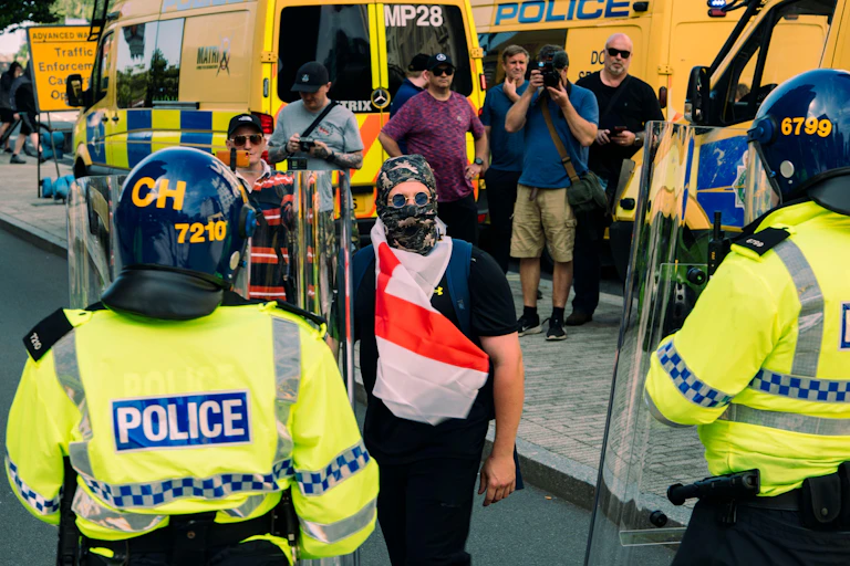Two riot police face to face with a man wearing an England flag and a camo balaclava, several police vans behind him