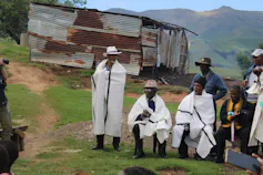 Six men stand in a rural area dressed in traditional clothing.