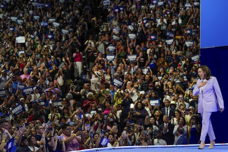 A dark-haired woman in a lilac-coloured pantsuit smiles as she walks onto a stage in front of thousands of people.
